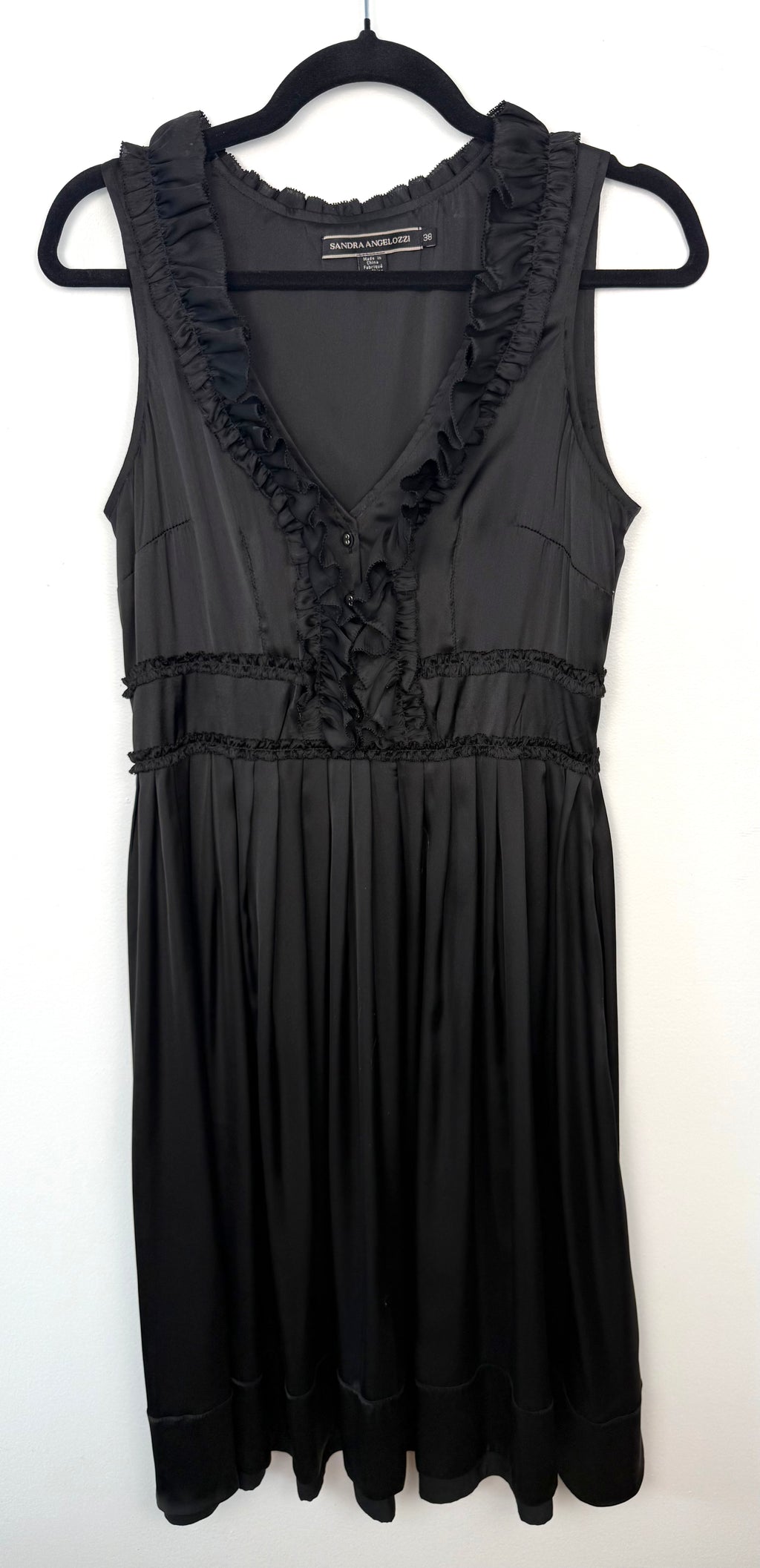 Black Sandra Angelozzi sleeveless dress with ruffle trim V-neckline and gathered waist, hanging on black hanger against white wall.