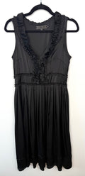 Black Sandra Angelozzi sleeveless dress with ruffle trim V-neckline and gathered waist, hanging on black hanger against white wall.
