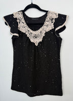 Black sequin knit top with cream crochet lace neckline hanging on black hanger