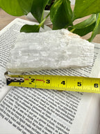 Crystal rock on top of a book with a measuring tape for scale, plant in the background. Measures approx 6"