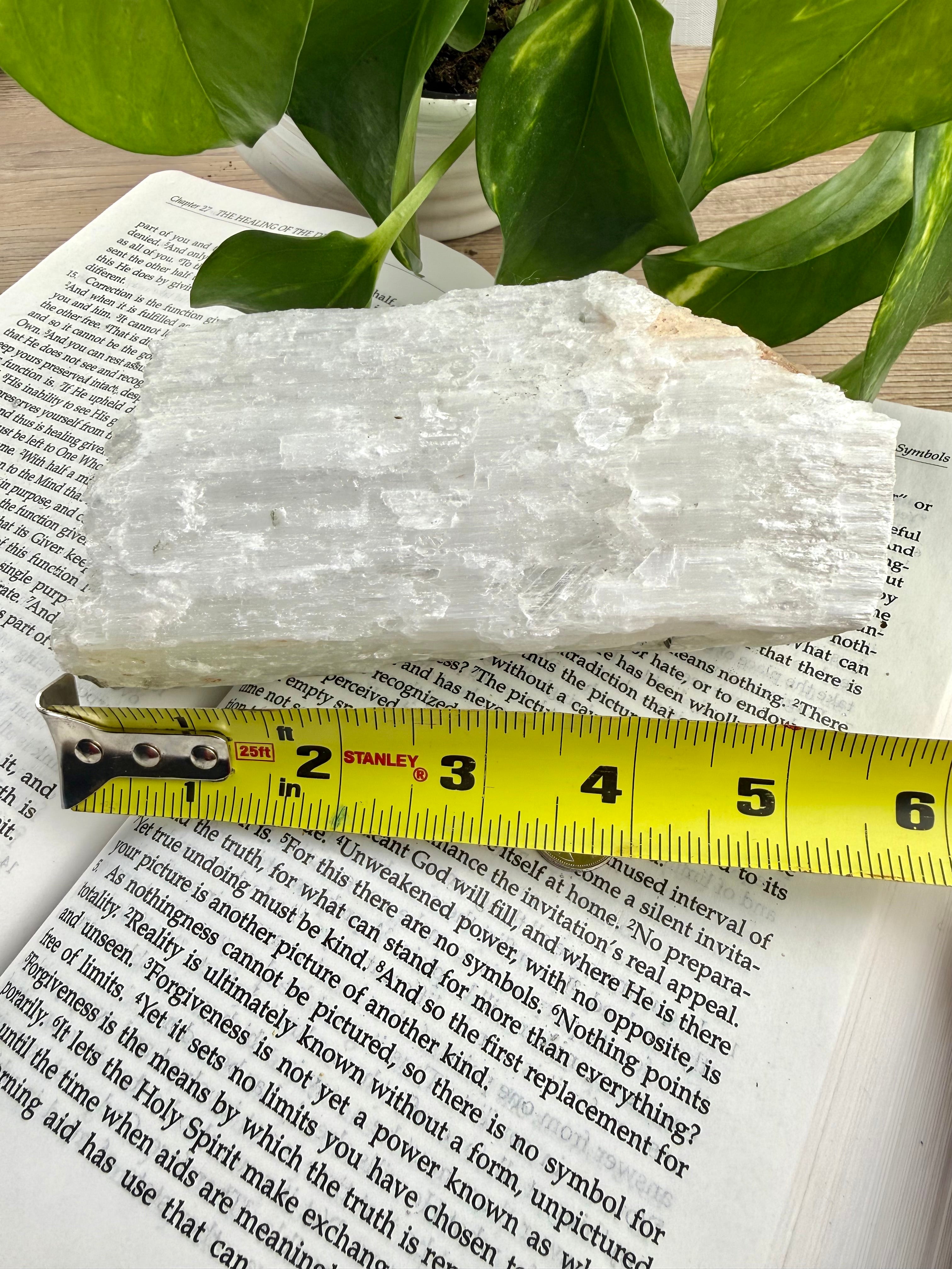 Crystal rock on top of a book with a measuring tape for scale, plant in the background. Measures approx 6"
